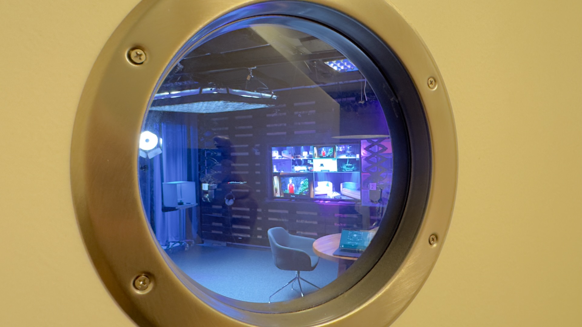 Blick durch das messingfarbene Bullaugen-Fenster ins Prismeo Studio mit Interview-Setup und Monitorwand