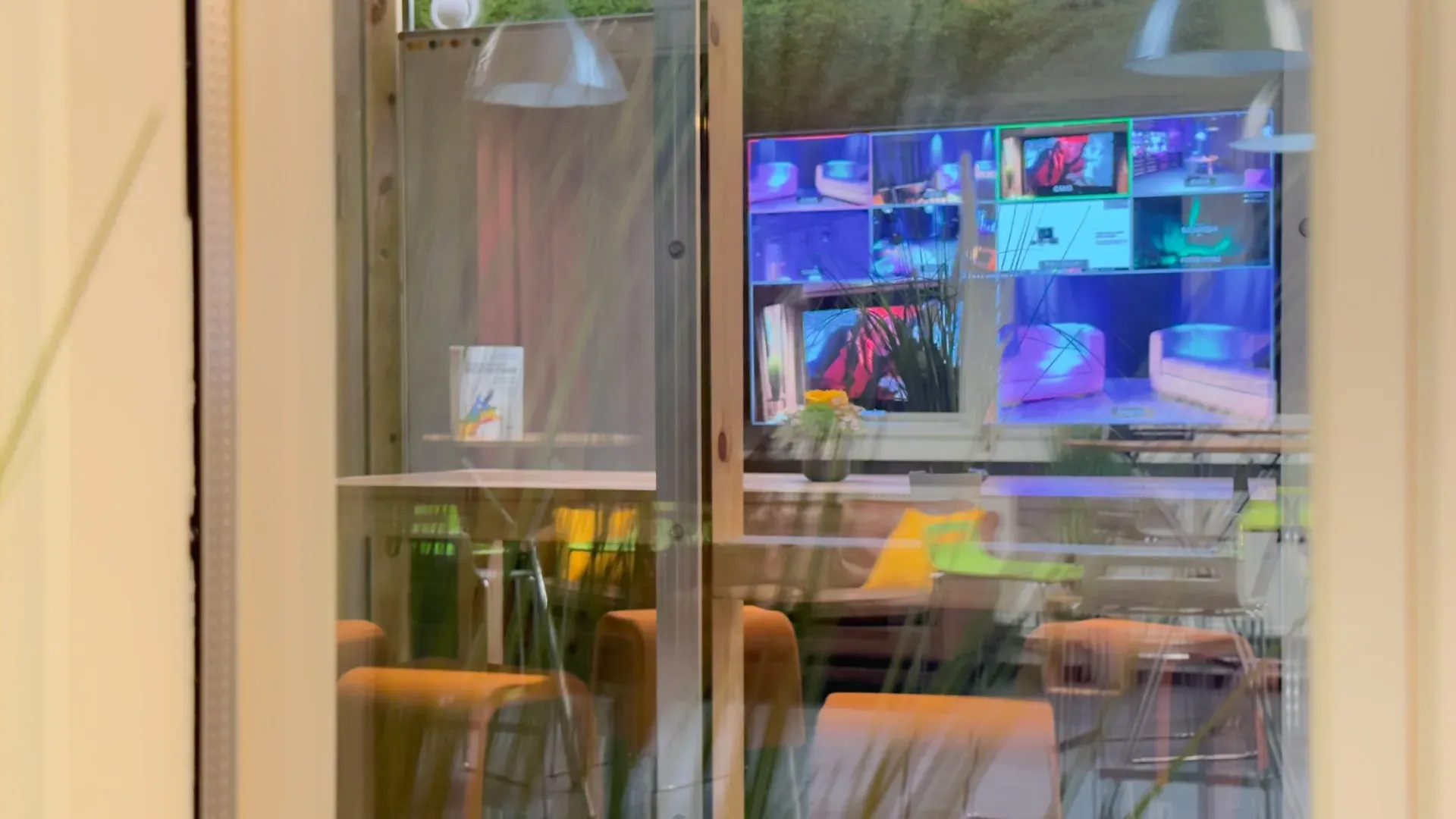 Greenhouse – Blick durch das Glasfenster ins Greenhouse mit Multi-View-Display und orangefarbenen Stühlen – Bezug zu den angrenzenden Räumen