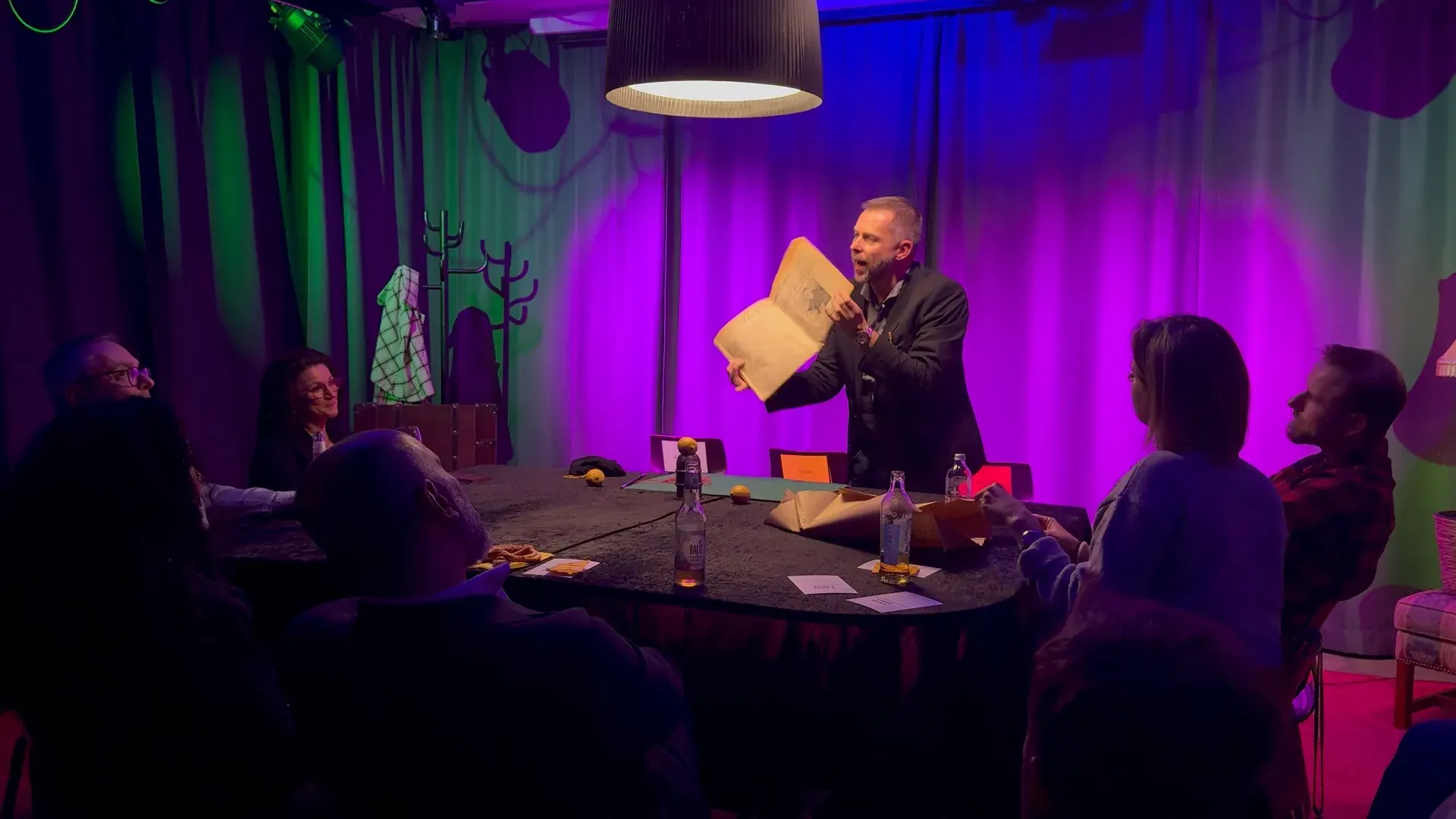 Theater – Zaubershow im Theater des Prismeo LAB mit Künstler am runden Tisch, altem Buch in der Hand und atmosphärischer lila-grüner Bühnenbeleuchtung