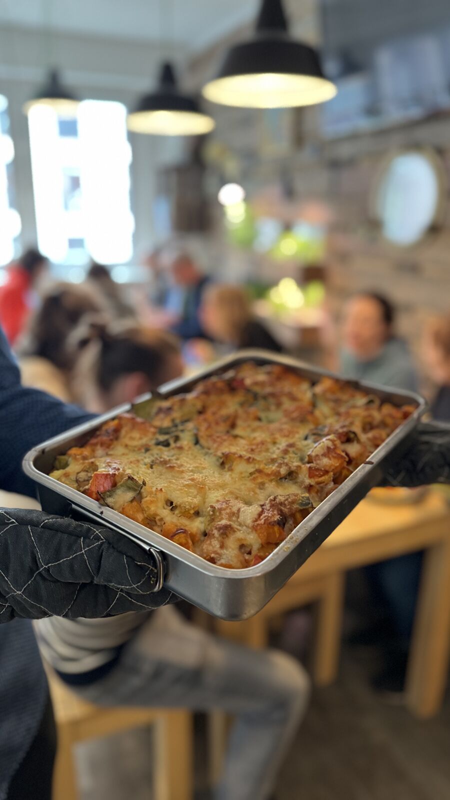 Eine Person hält eine Auflaufform mit überbackenem Essen, im Hintergrund verschwommen Menschen.