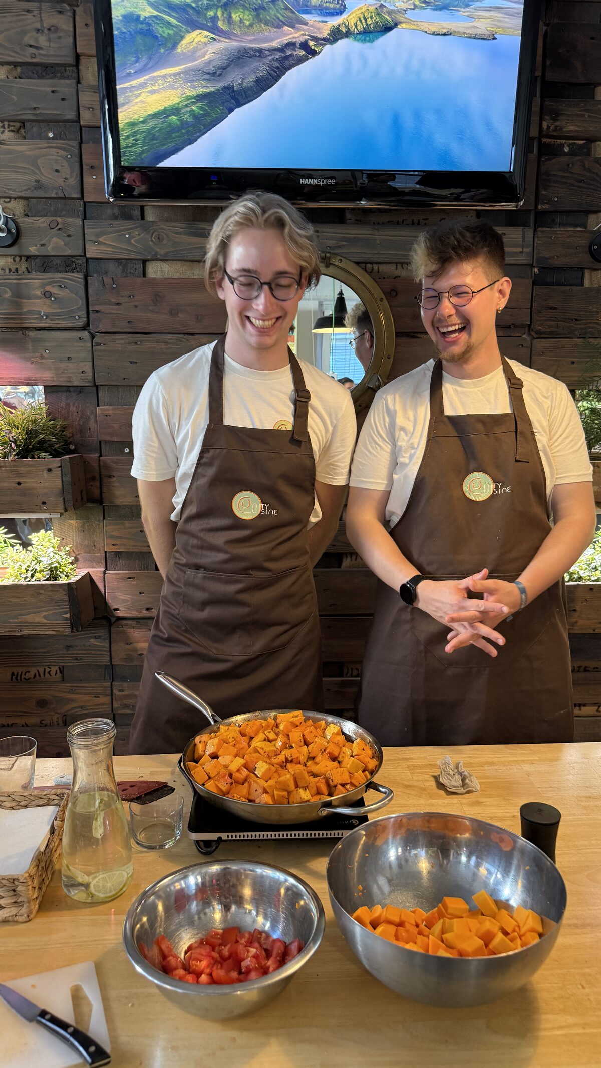 Zwei lachende junge Köche in Schürzen hinter einer Pfanne mit gerösteten Kürbiswürfeln und Schalen mit Tomaten in der Hongkong Kitchen.